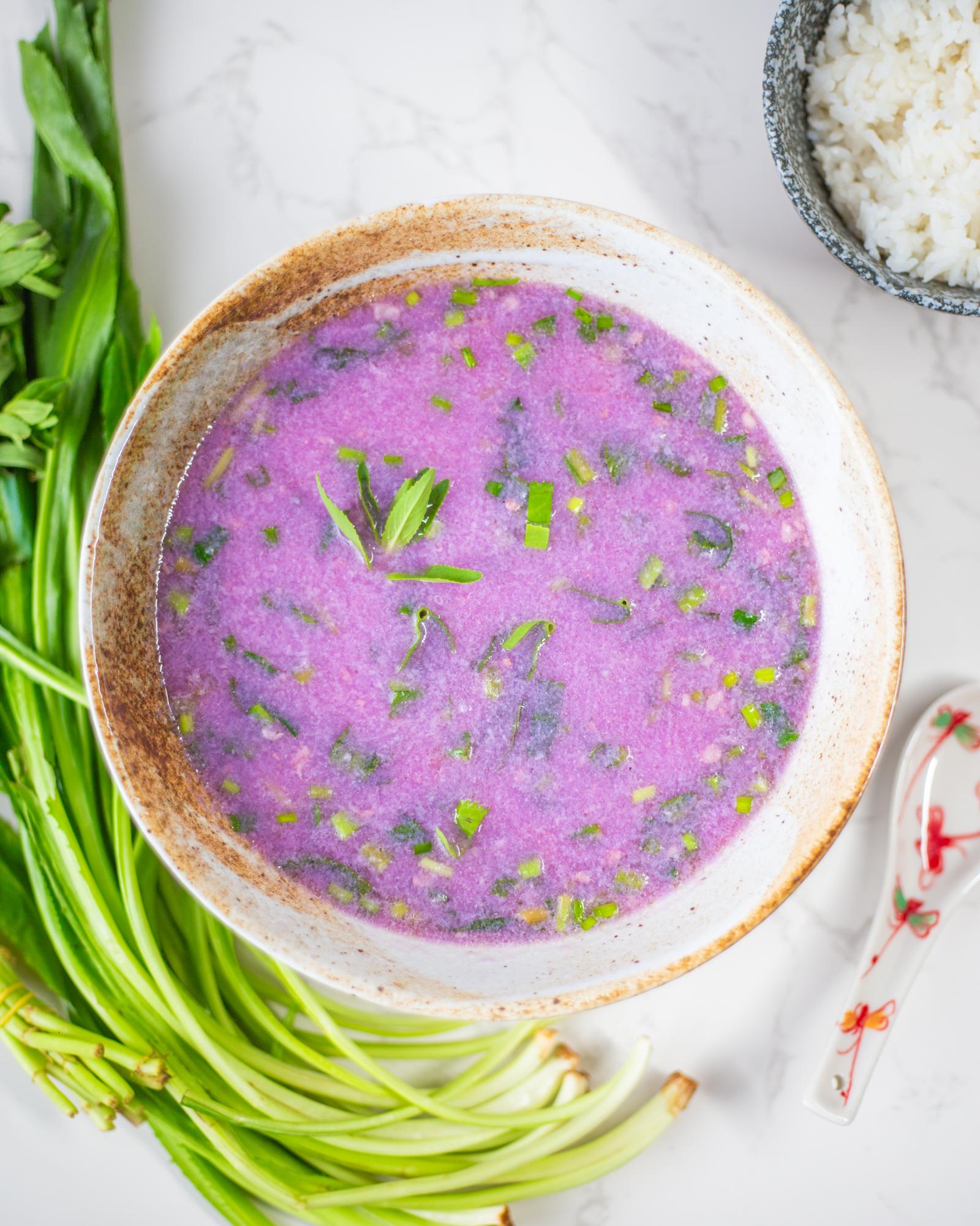 A bowl filled with a bright purple soup made with purple yam, along with chopped herbs. There are bundles of herbs next to the bowl, as well as a small bowl of rice and a soup spoon