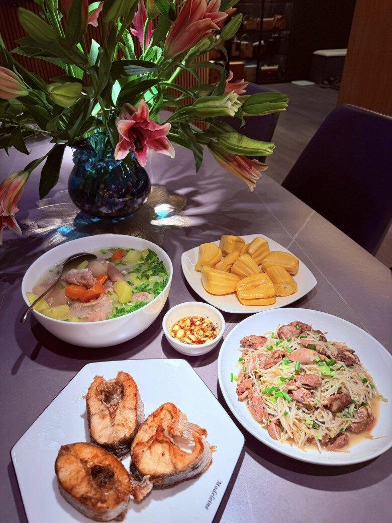 Mâm cơm đầy đủ dưỡng chất với các món: Canh xương ninh khoai tây - cà rốt, cá chiên vàng ruộm, tim gà xào giá, nước chấm tỏi ớt pha đậm đà và mít tráng miệng. Không gian ăn uống thêm ấm cúng với bình hoa ly rực rỡ, đúng như lời chia sẻ của Minh Hương: “Hạnh phúc đôi khi chỉ là căn bếp ấm và nồi cơm đang sôi…”